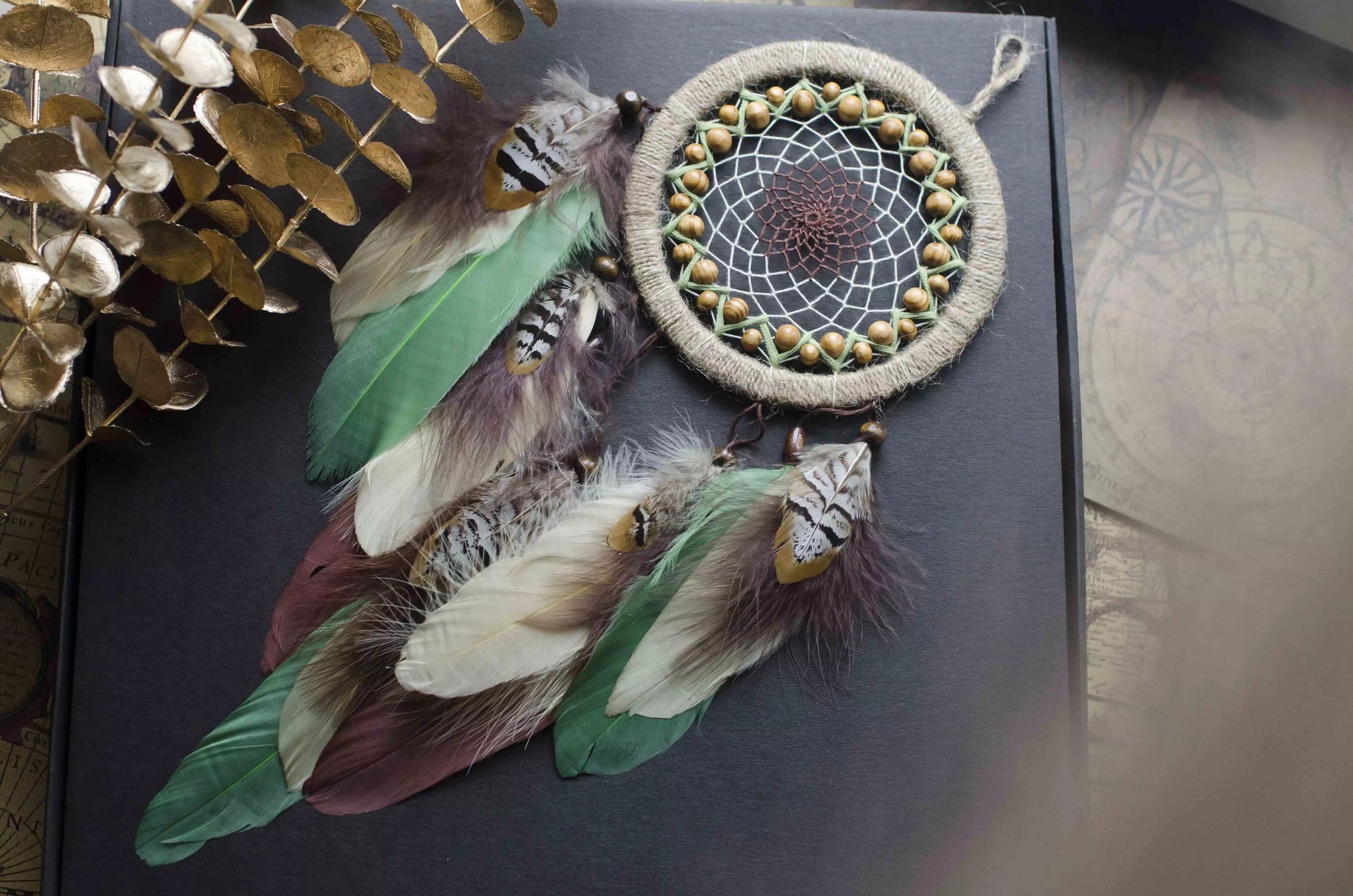 Handmade dream catcher with a 6-inch hoop wrapped in natural jute twine, featuring an intricate web design with wooden beads. Adorned with an array of feathers in green, cream, and brown tones, it serves as a beautiful boho wall decor piece. Perfect for adding a touch of rustic charm to any room.