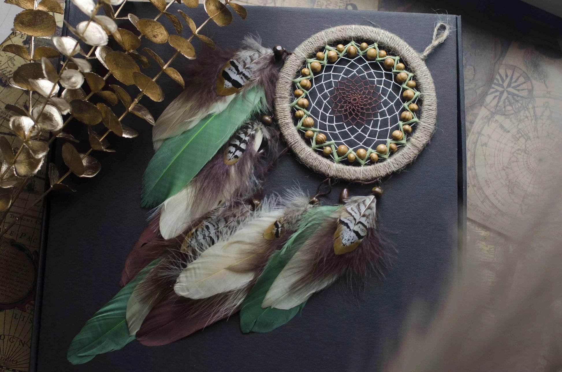 Handmade dream catcher with a 6-inch hoop wrapped in natural jute twine, featuring an intricate web design with wooden beads. Adorned with an array of feathers in green, cream, and brown tones, it serves as a beautiful boho wall decor piece. Perfect for adding a touch of rustic charm to any room.