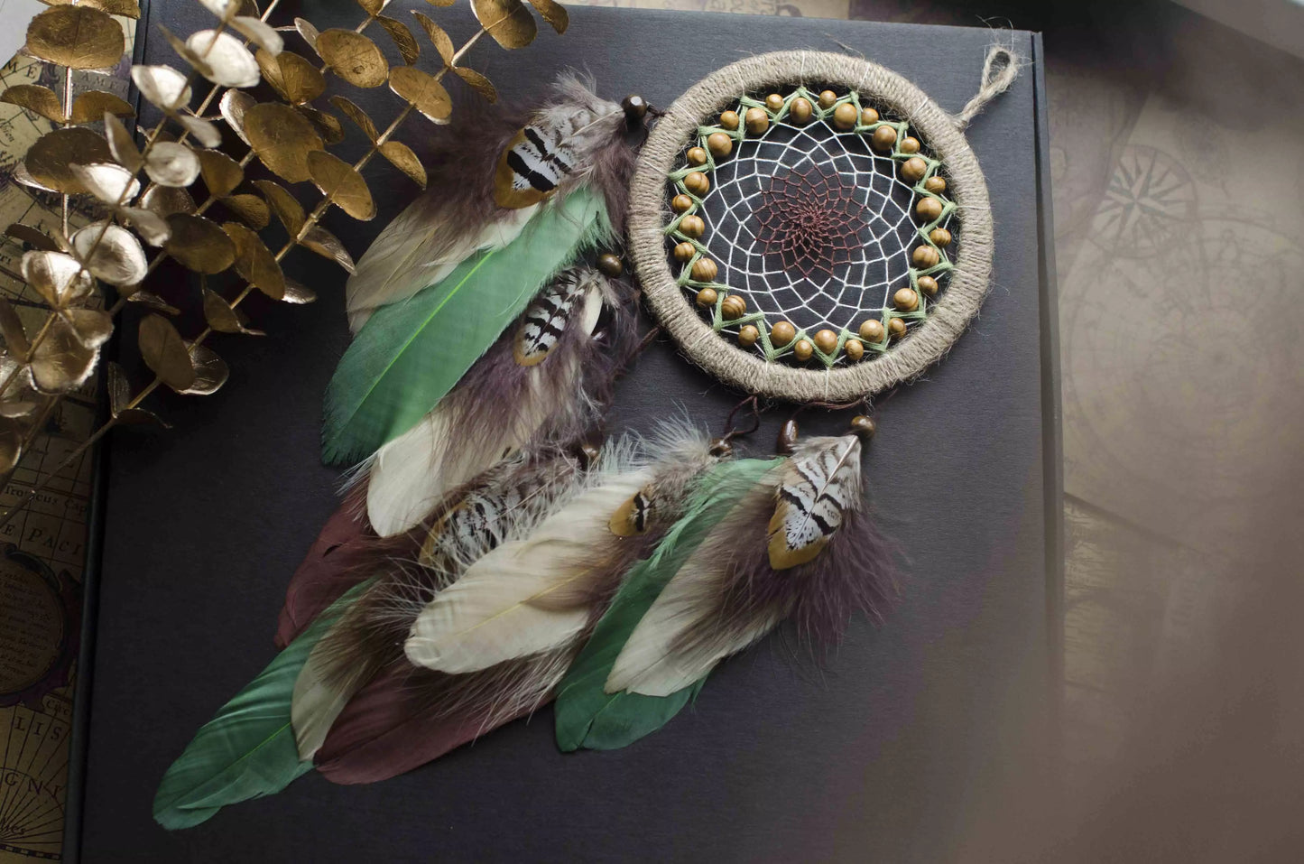 Handmade dream catcher with a 6-inch hoop wrapped in natural jute twine, featuring an intricate web design with wooden beads. Adorned with an array of feathers in green, cream, and brown tones, it serves as a beautiful boho wall decor piece. Perfect for adding a touch of rustic charm to any room.