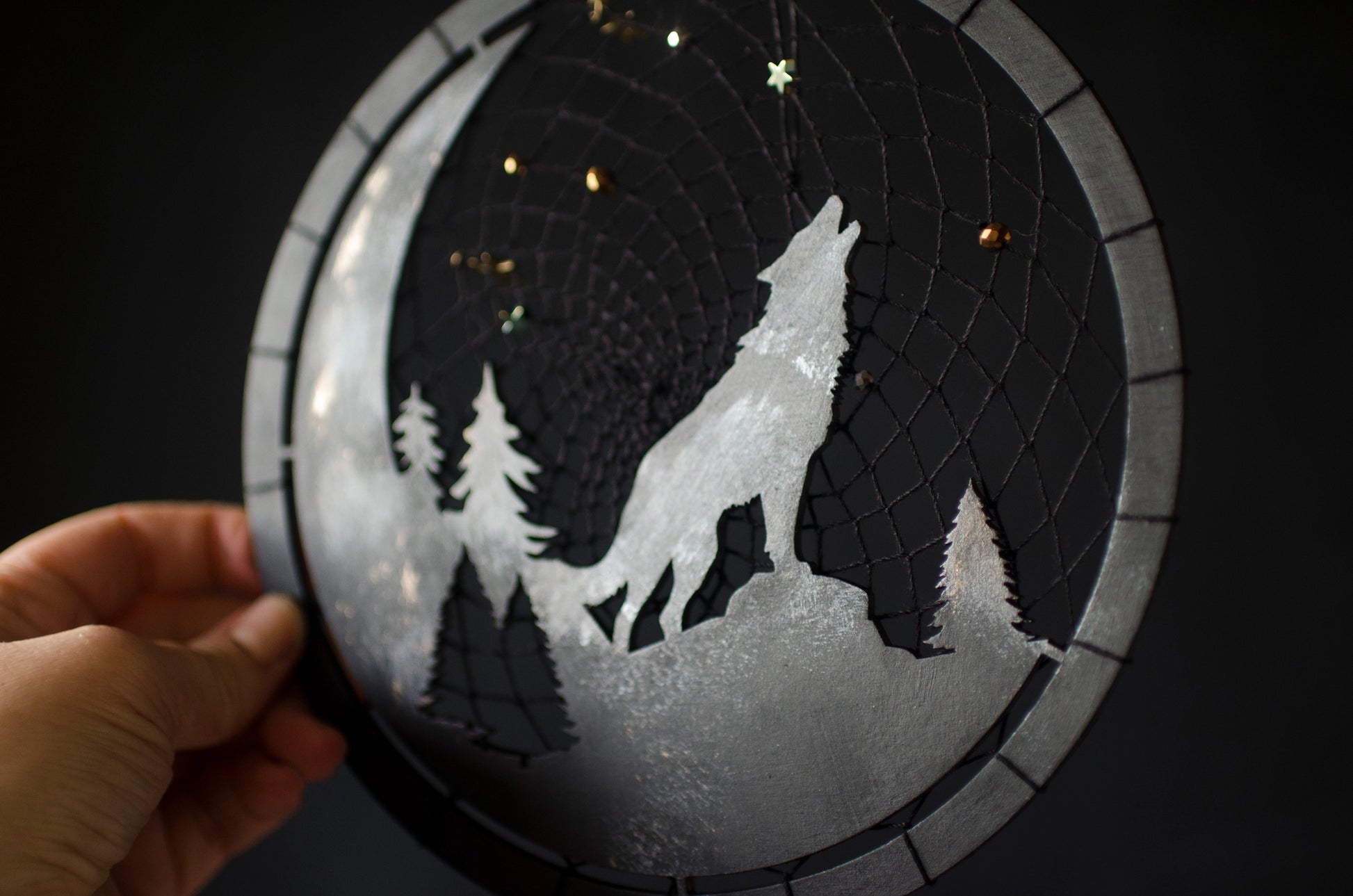 A handcrafted dream catcher with a howling wolf silhouette, moon, and forest design. Decorated with star-shaped beads, this unique piece captures the essence of the night. The hoop measures approximately 8 inches in diameter, featuring black and natural shades.