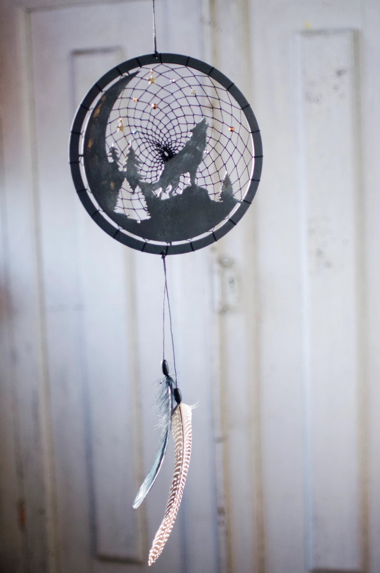 A handcrafted dream catcher with a howling wolf silhouette, moon, and forest design. Decorated with star-shaped beads, this unique piece captures the essence of the night. The hoop measures approximately 8 inches in diameter, featuring black and natural shades.