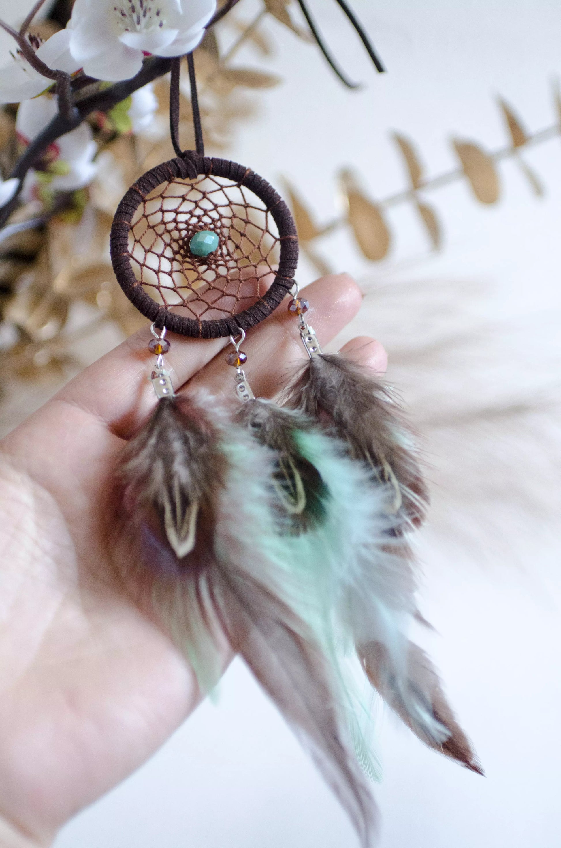 Handmade small dreamcatcher featuring a brown web, turquoise stone center, and brown and mint green feathers, perfect as boho car decor or for adding positive energy and protection to any space.