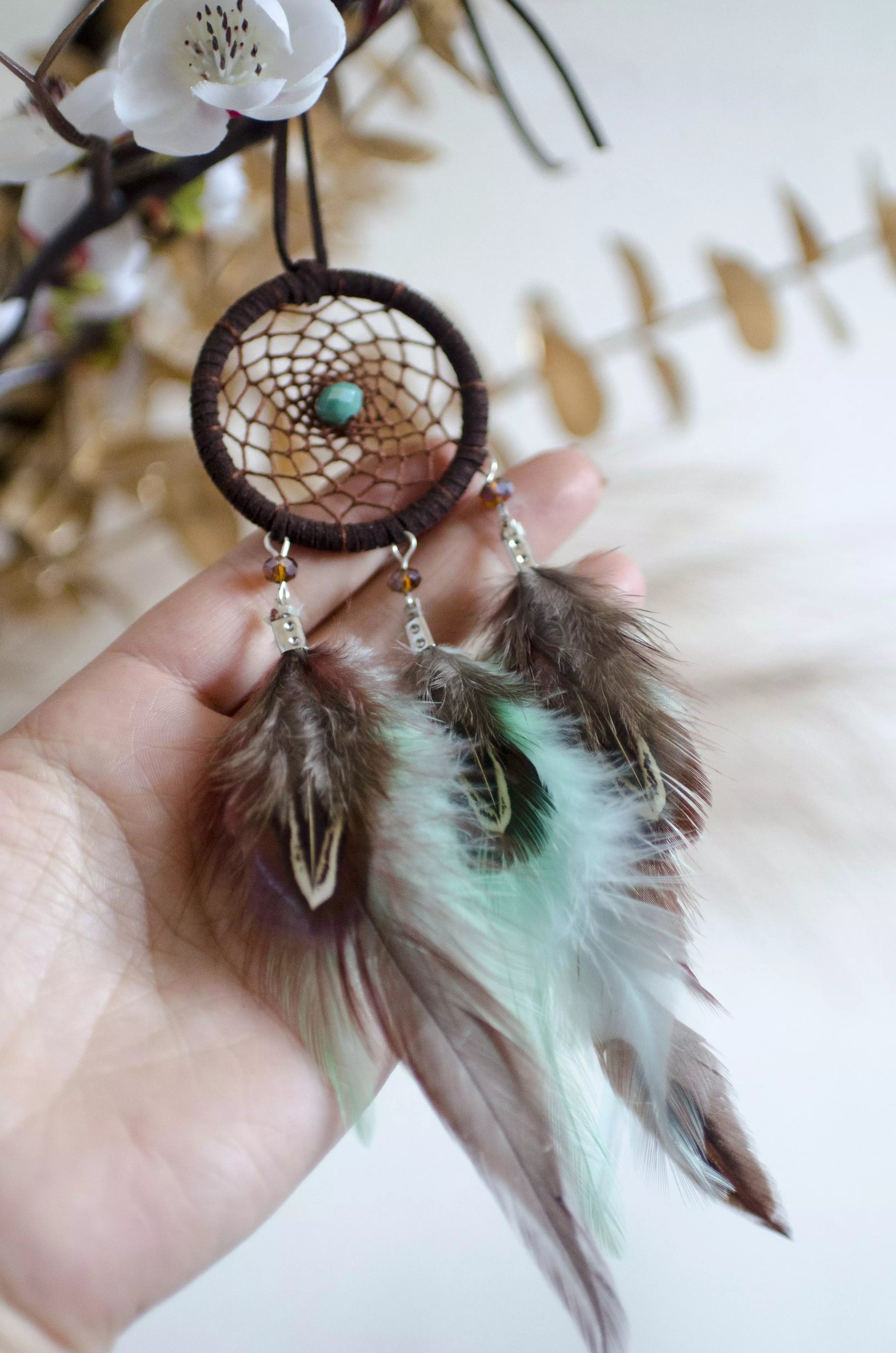 Handmade small dreamcatcher featuring a brown web, turquoise stone center, and brown and mint green feathers, perfect as boho car decor or for adding positive energy and protection to any space.