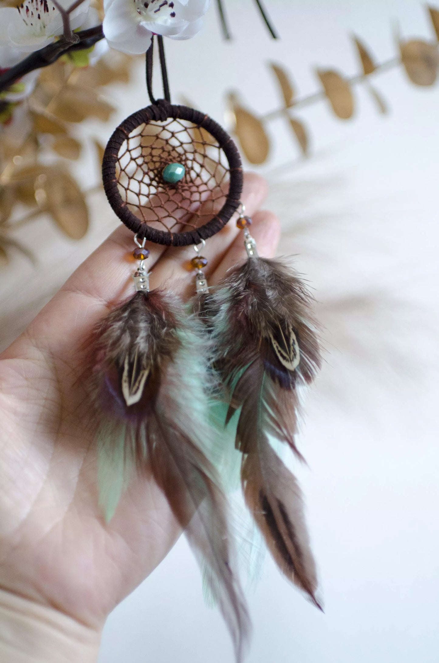 Handmade small dreamcatcher featuring a brown web, turquoise stone center, and brown and mint green feathers, perfect as boho car decor or for adding positive energy and protection to any space.