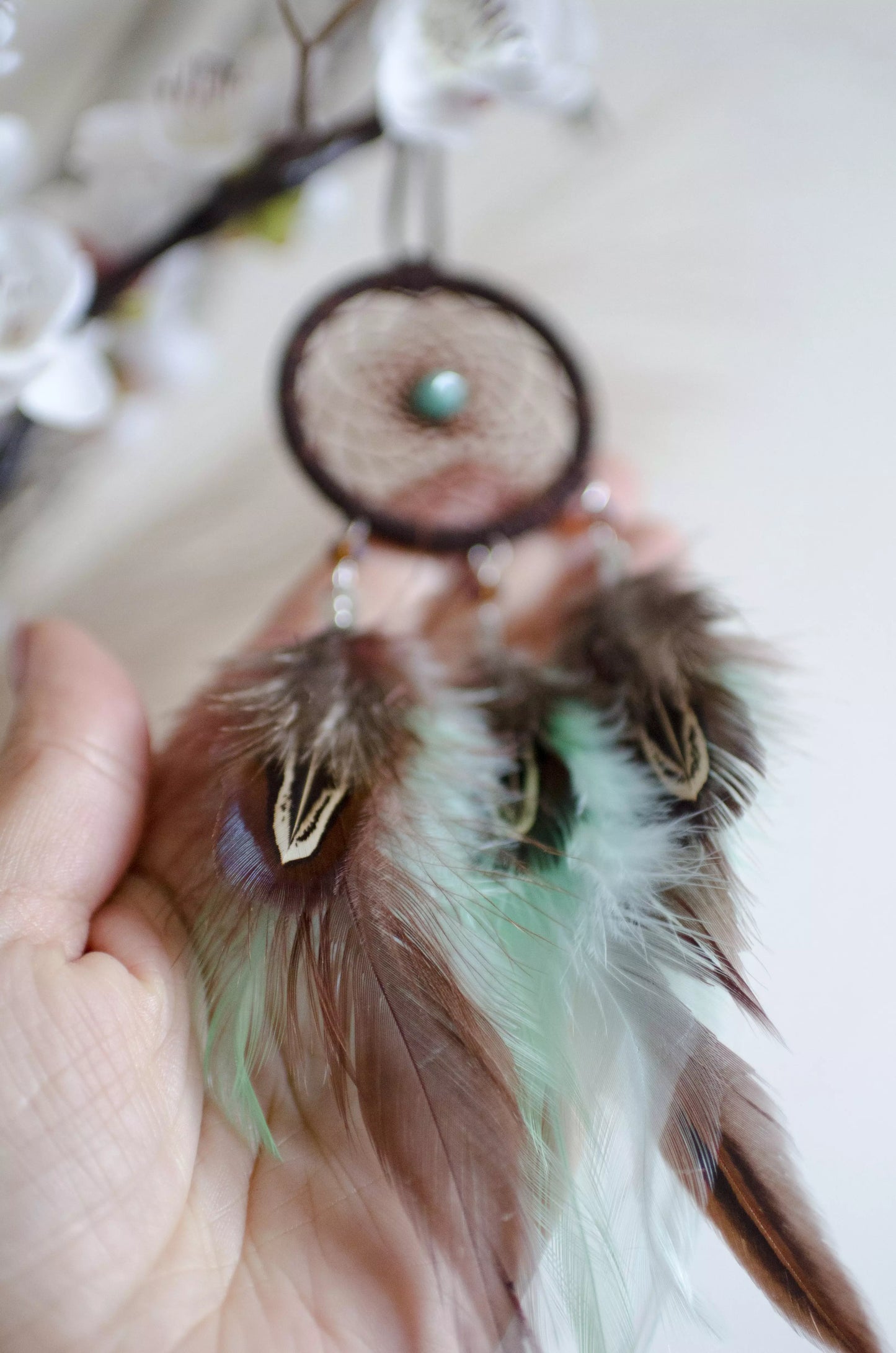 Handmade small dreamcatcher featuring a brown web, turquoise stone center, and brown and mint green feathers, perfect as boho car decor or for adding positive energy and protection to any space.