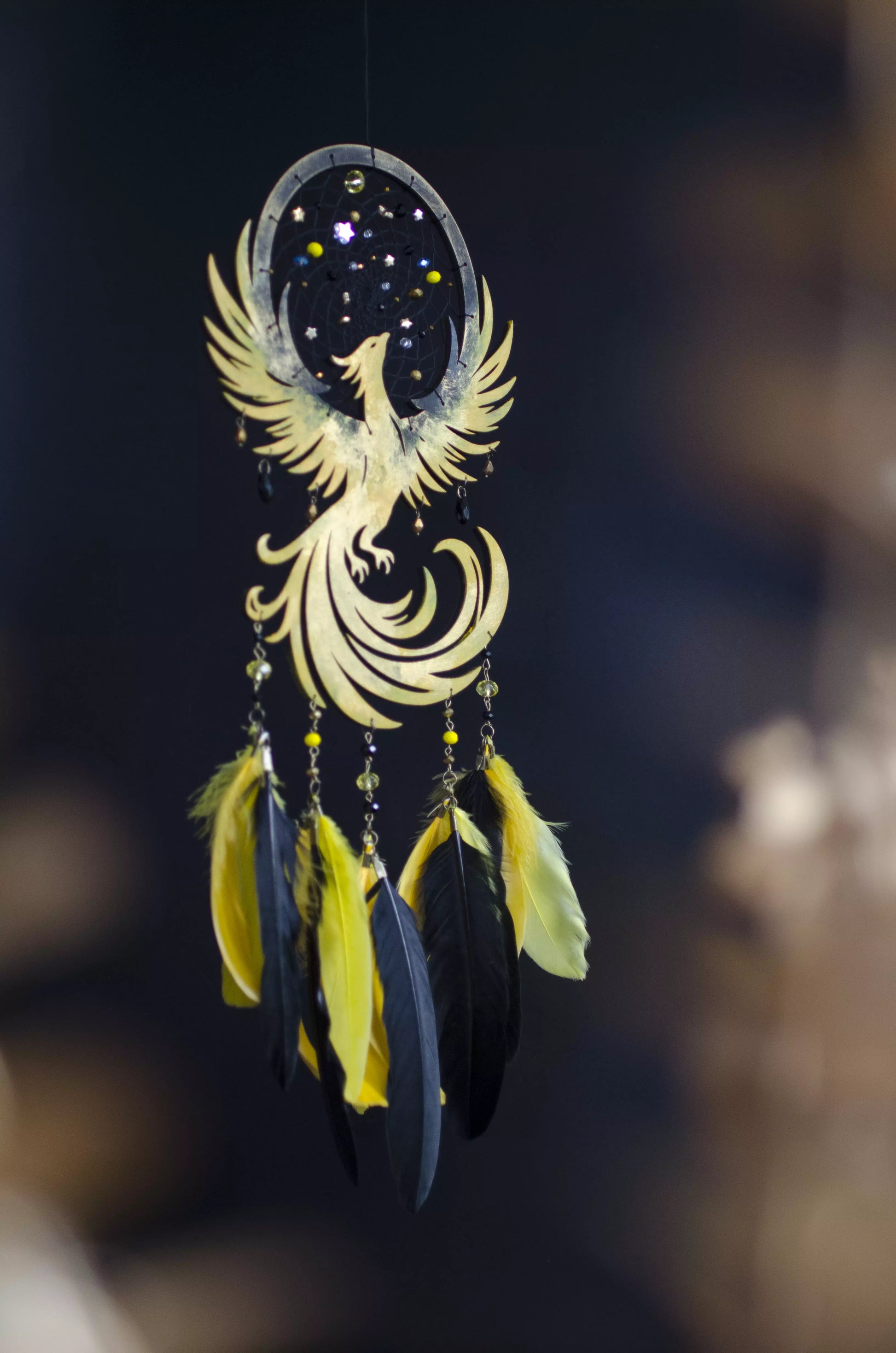 Handmade golden phoenix dreamcatcher with a celestial black web adorned with star and bead accents, featuring a soaring phoenix silhouette and strands decorated with bright yellow and deep black feathers, set against a softly blurred dark background.