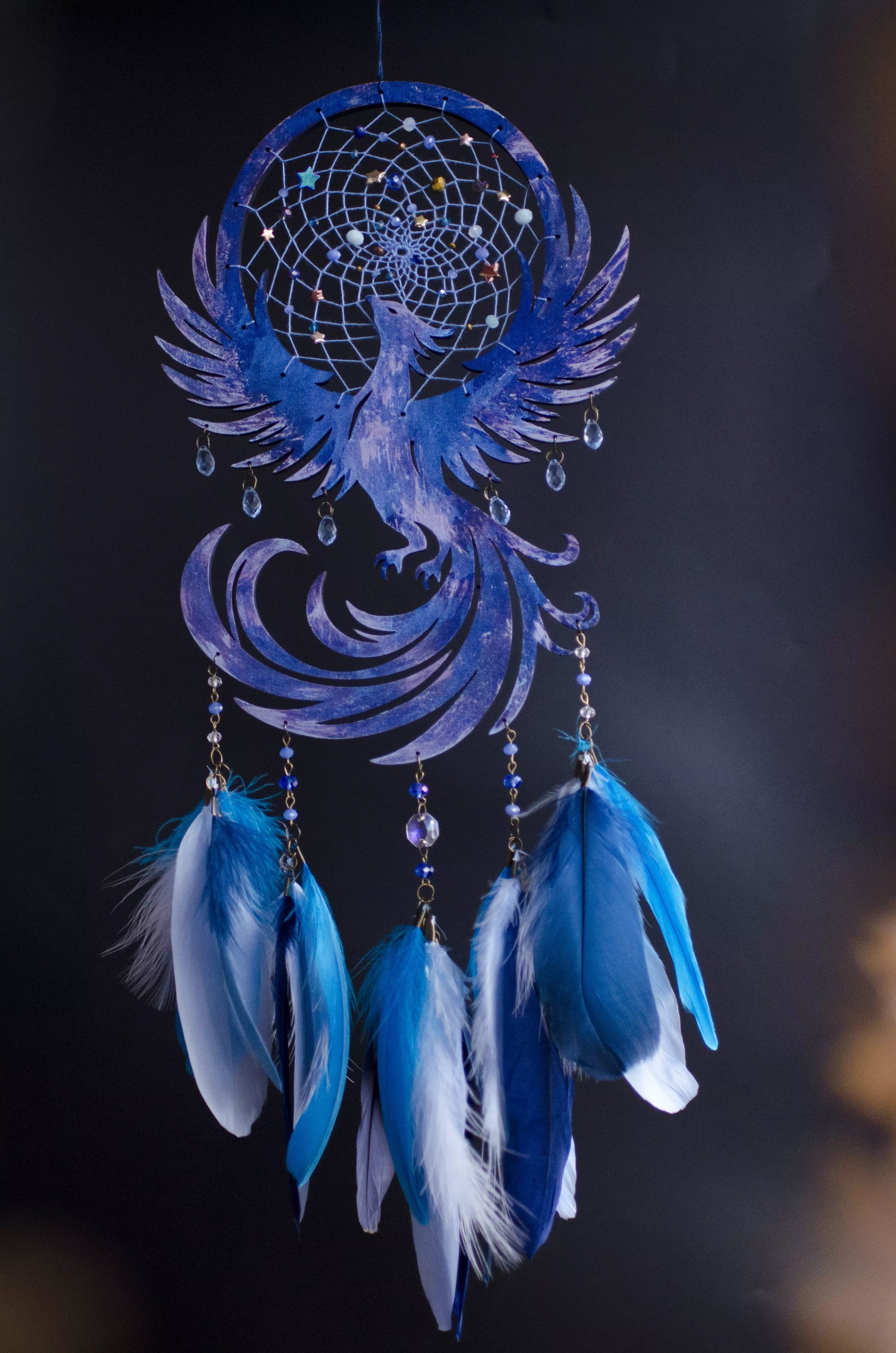 Handcrafted blue and purple phoenix dreamcatcher featuring an intricate web with celestial bead accents, a soaring phoenix silhouette at the center, and long strands adorned with vibrant navy and light blue feathers, hanging elegantly against a dark