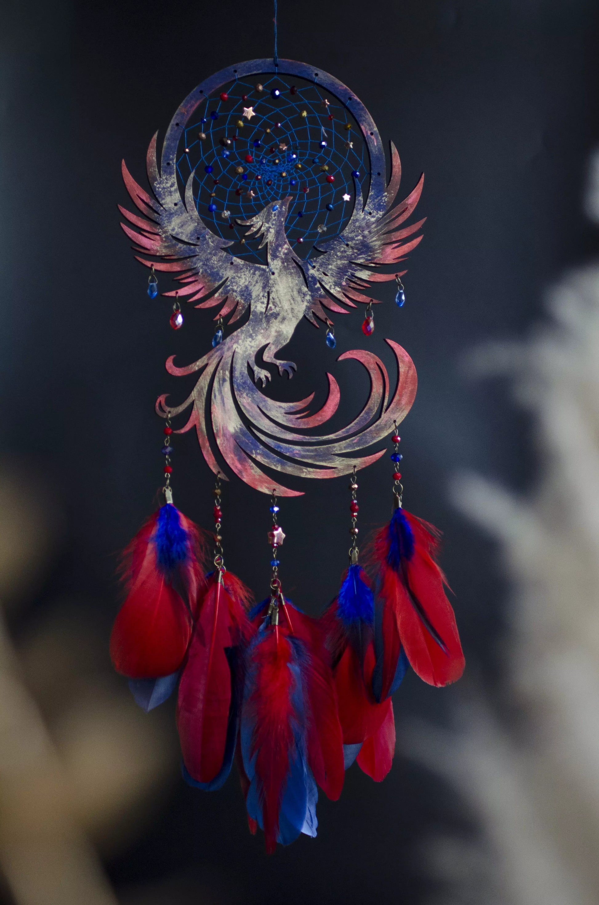 Handcrafted phoenix dreamcatcher with a detailed blue web adorned with celestial bead accents, featuring a soaring phoenix silhouette and strands decorated with vibrant red and deep blue feathers, hanging gracefully against a dark, softly blurred background.