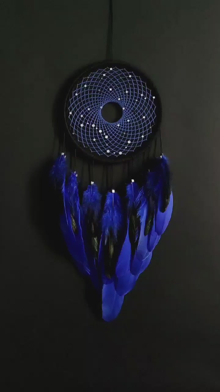 Handmade dreamcatcher with a black hoop, featuring a meticulously woven web adorned with small reflective beads, creating a galaxy-like effect. The dreamcatcher is embellished with vibrant blue and purple feathers, adding a striking contrast and visual appeal. Perfect for adding a touch of mystique and elegance to any space.