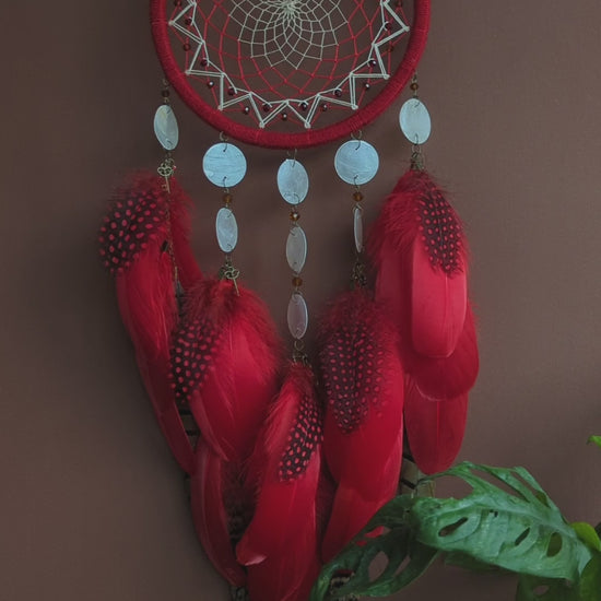 Red dream catcher with a detailed white web, adorned with white beads and vibrant red feathers. Perfect for adding a touch of boho charm and positive energy to your space. Hangs elegantly against a backdrop of greenery, creating a striking visual contrast.