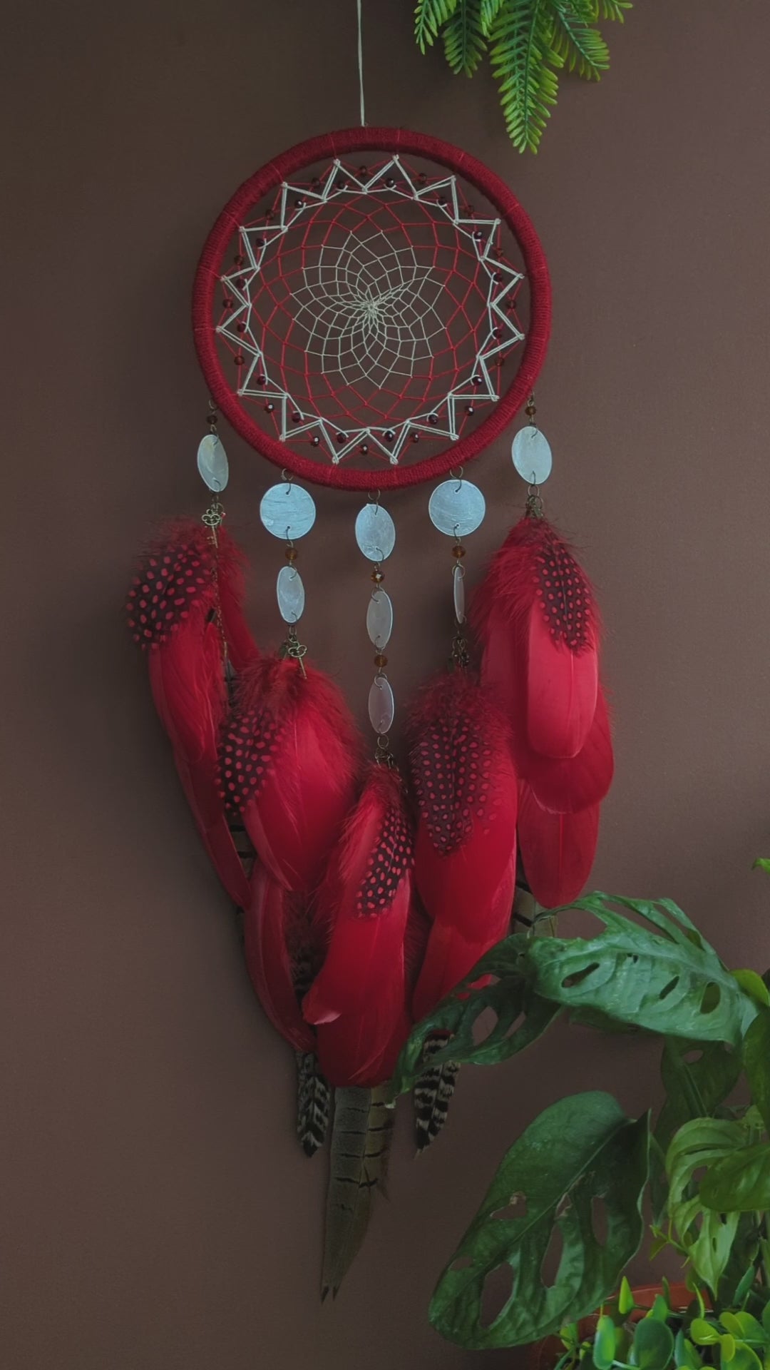 Red dream catcher with a detailed white web, adorned with white beads and vibrant red feathers. Perfect for adding a touch of boho charm and positive energy to your space. Hangs elegantly against a backdrop of greenery, creating a striking visual contrast.