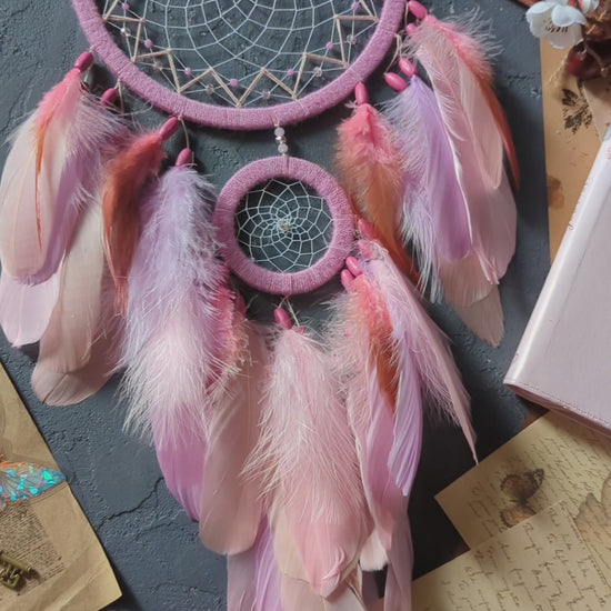 This dream catcher features a large pink hoop with an intricate web adorned with pink beads. Below, it displays an array of soft pink and orange feathers, accented with additional pink beads. The design is complemented by surrounding vintage letters, keys, and delicate cherry blossoms, creating a charming and whimsical aesthetic perfect for any space.