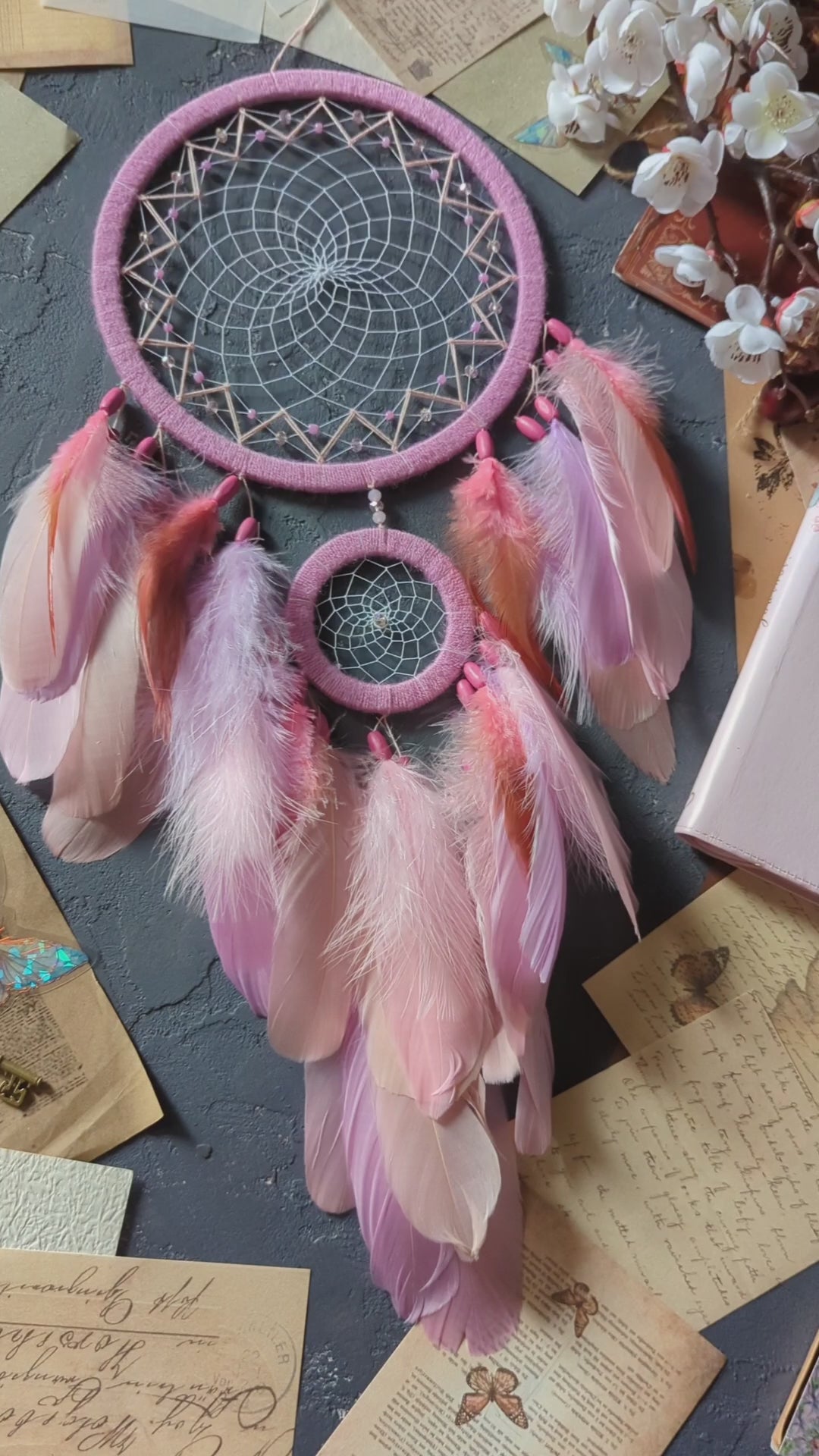 This dream catcher features a large pink hoop with an intricate web adorned with pink beads. Below, it displays an array of soft pink and orange feathers, accented with additional pink beads. The design is complemented by surrounding vintage letters, keys, and delicate cherry blossoms, creating a charming and whimsical aesthetic perfect for any space.