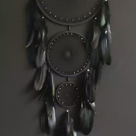 Handcrafted dream catcher featuring a striking black design, with three concentric hoops of varying sizes adorned with intricate webbing. Each hoop is decorated with black beads and complemented by an array of natural black feathers. This elegant piece measures a total length that ensures it becomes a captivating focal point in any room. Perfect for adding a touch of sophistication and positive energy to your home decor. Ideal for those who appreciate unique, artisan-made creations.