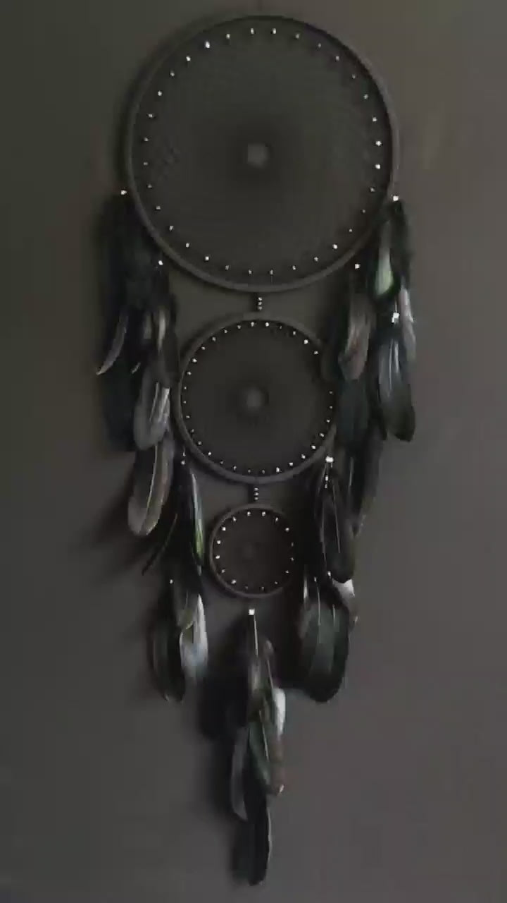 Handcrafted dream catcher featuring a striking black design, with three concentric hoops of varying sizes adorned with intricate webbing. Each hoop is decorated with black beads and complemented by an array of natural black feathers. This elegant piece measures a total length that ensures it becomes a captivating focal point in any room. Perfect for adding a touch of sophistication and positive energy to your home decor. Ideal for those who appreciate unique, artisan-made creations.