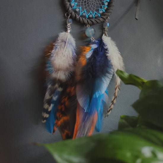 Small dreamcatcher for car in shades of gray, turquoise, blue, and orange. Features intricate webbing with a central turquoise bead, surrounded by feathers and beads in coordinating colors. Perfect for adding a touch of boho charm to your vehicle