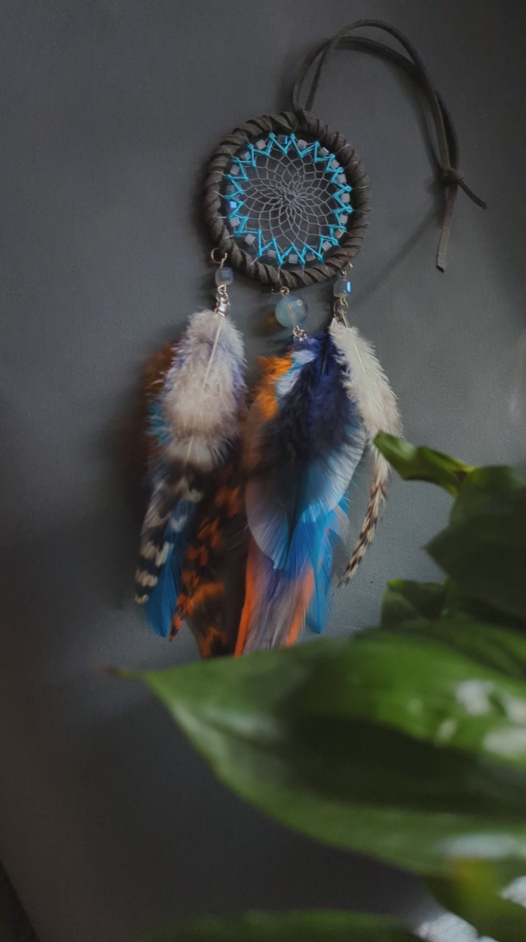 Small dreamcatcher for car in shades of gray, turquoise, blue, and orange. Features intricate webbing with a central turquoise bead, surrounded by feathers and beads in coordinating colors. Perfect for adding a touch of boho charm to your vehicle