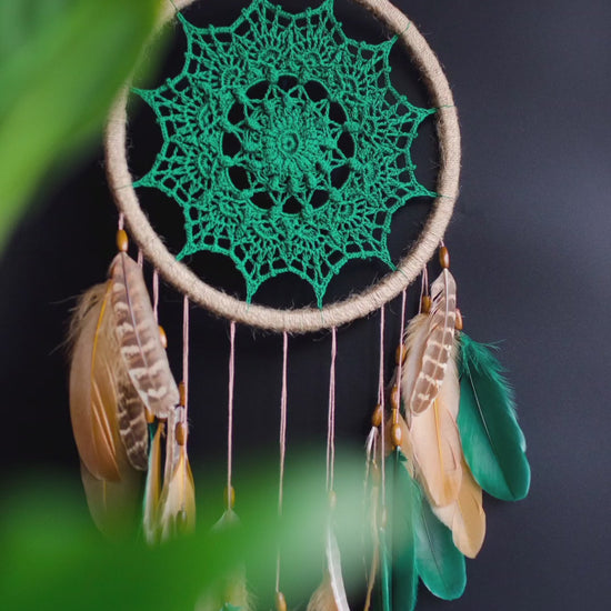 A handcrafted dreamcatcher featuring a green crochet mandala centerpiece, wrapped in a jute hoop with natural brown and green feathers hanging below, styled against dark and light backgrounds for a modern, bohemian look.