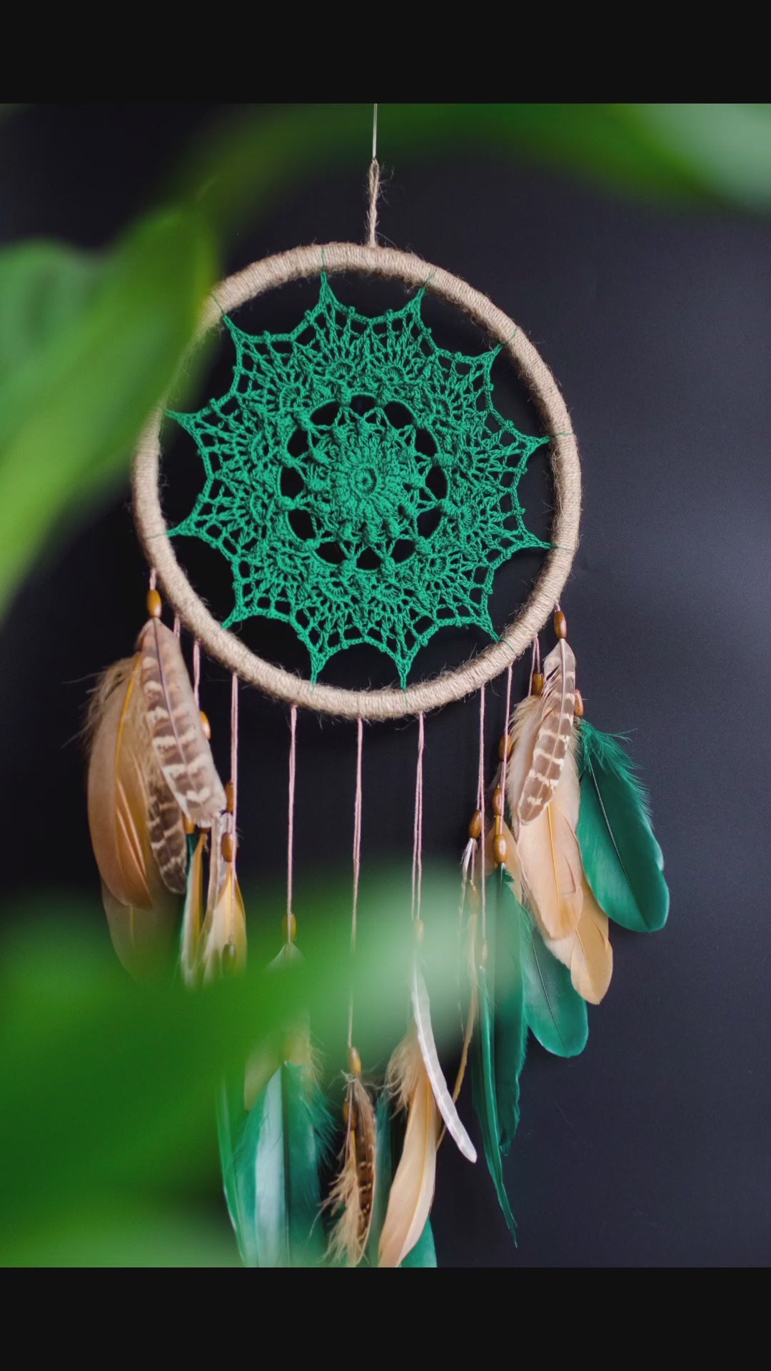 A handcrafted dreamcatcher featuring a green crochet mandala centerpiece, wrapped in a jute hoop with natural brown and green feathers hanging below, styled against dark and light backgrounds for a modern, bohemian look.