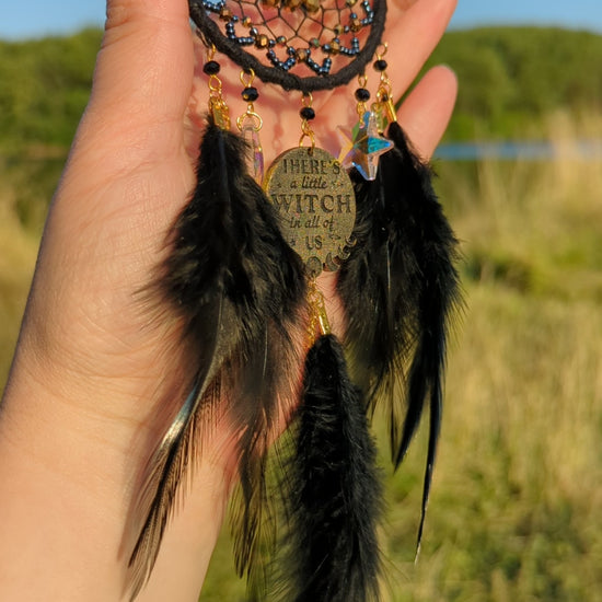 Handcrafted black witch dream catcher featuring a woven black yarn web, golden beads, black feathers, crystal moon and star charms, and a metallic 'There's a little witch in all of us' charm, designed for mystical decor and spiritual protection.