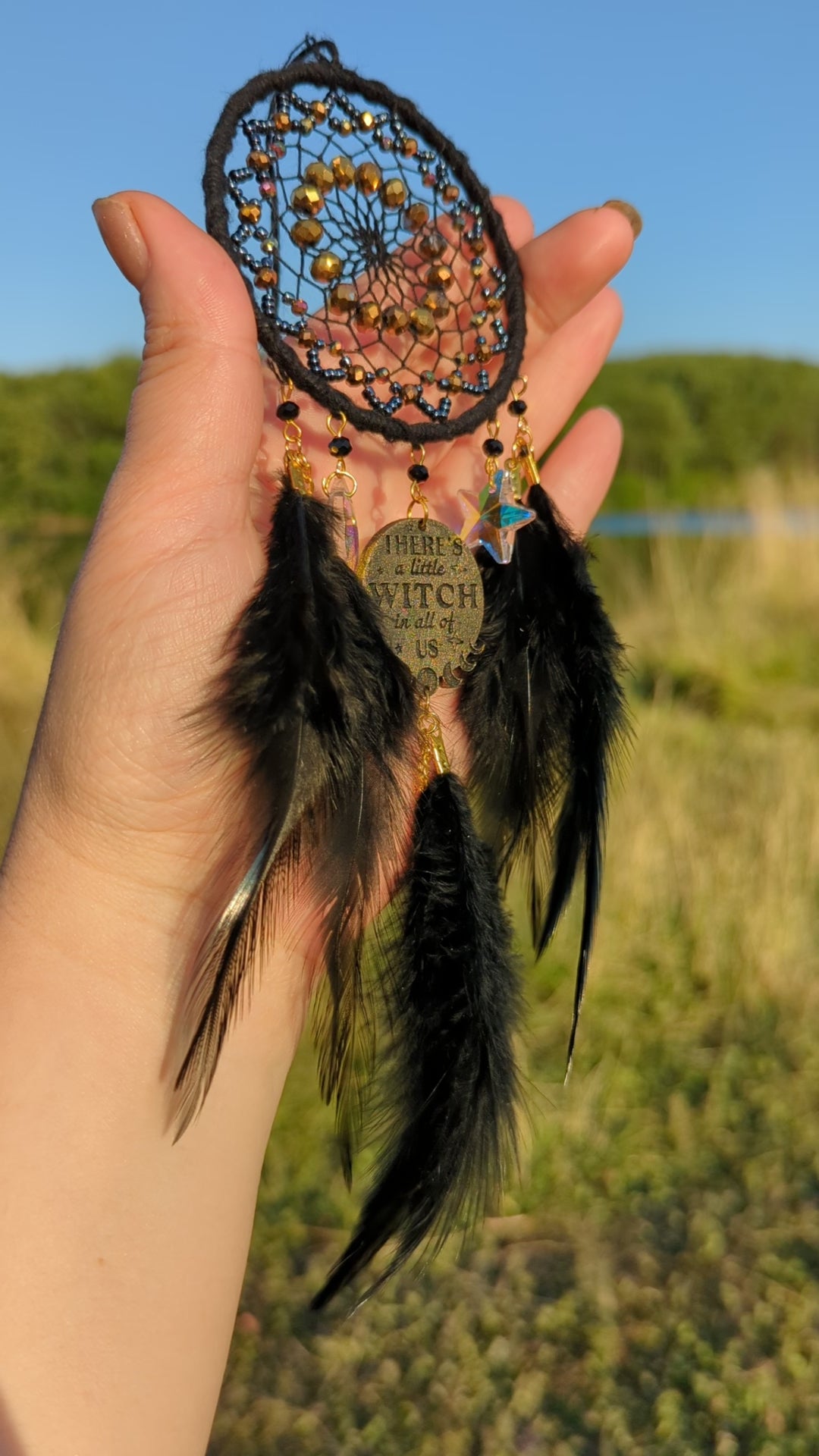 Handcrafted black witch dream catcher featuring a woven black yarn web, golden beads, black feathers, crystal moon and star charms, and a metallic 'There's a little witch in all of us' charm, designed for mystical decor and spiritual protection.