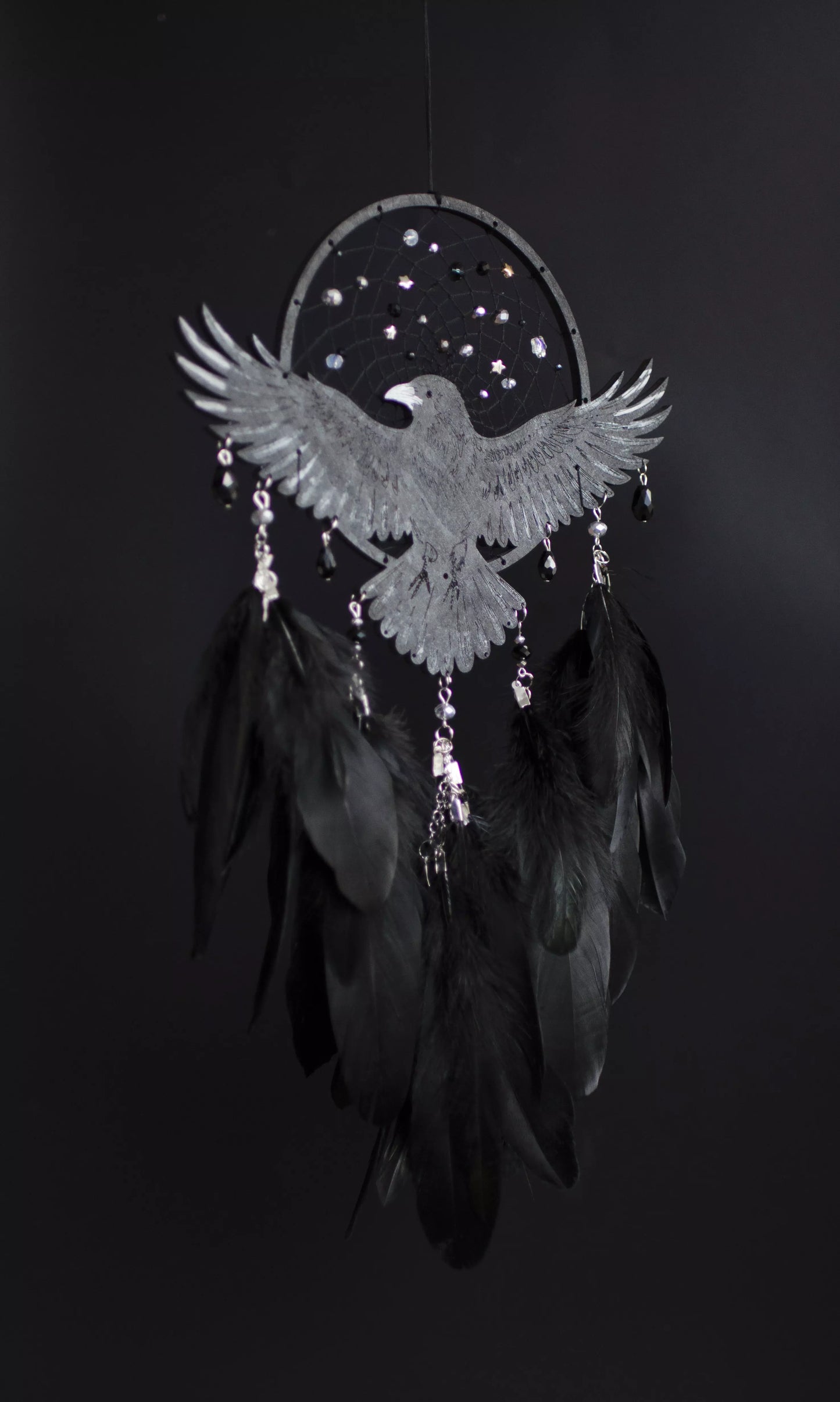 Handmade raven dream catcher with outstretched wings, set on a black hoop with a delicate web decorated with small reflective beads. Black feathers hang below with subtle crystal accents. Dark aesthetic wall hanging in charcoal and silver tones, detailed and symbolic design, perfect for moody interiors or meaningful gift.