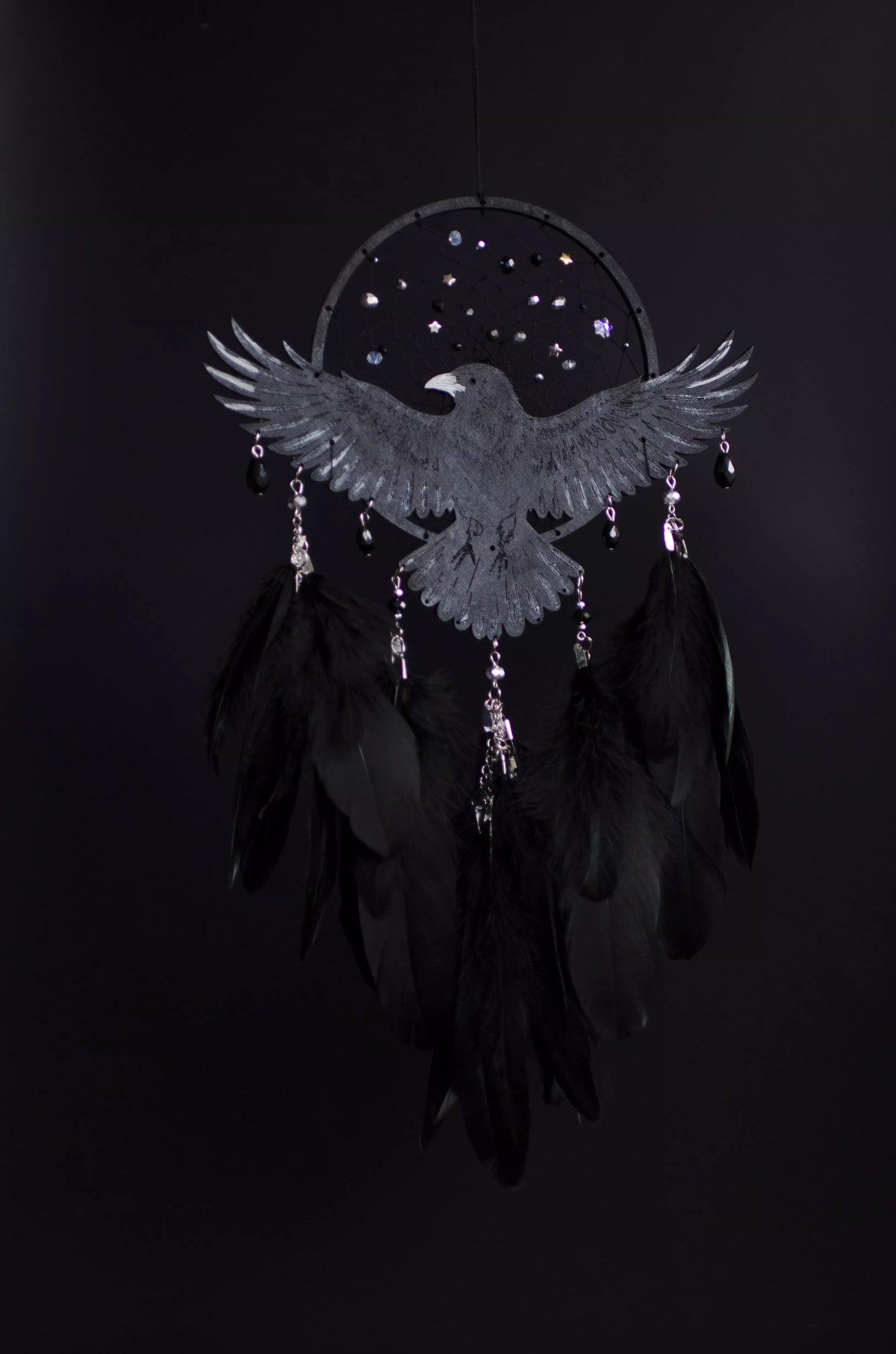 Handmade raven dream catcher with outstretched wings, set on a black hoop with a delicate web decorated with small reflective beads. Black feathers hang below with subtle crystal accents. Dark aesthetic wall hanging in charcoal and silver tones, detailed and symbolic design, perfect for moody interiors or meaningful gift.