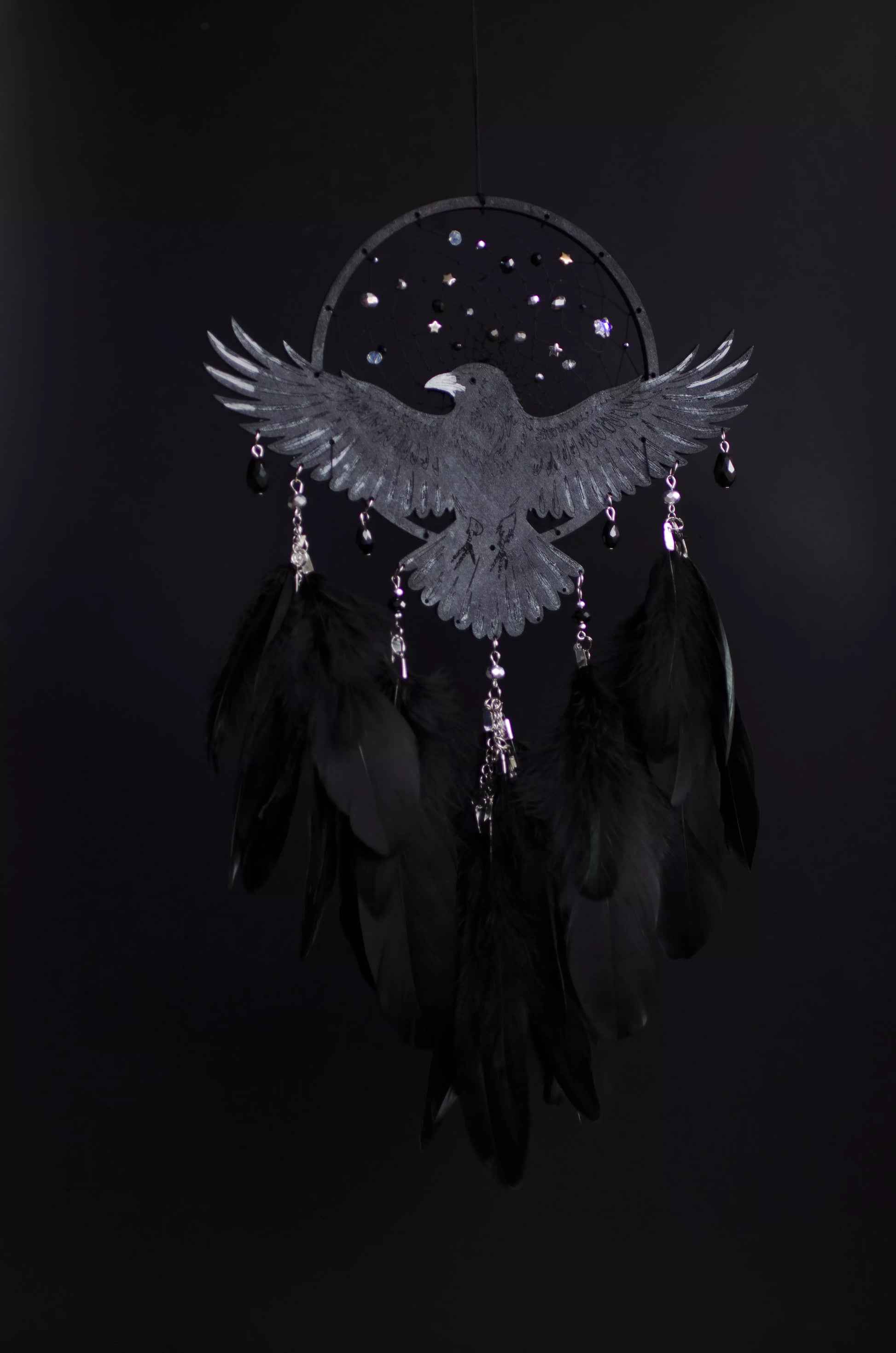 Handmade raven dream catcher with outstretched wings, set on a black hoop with a delicate web decorated with small reflective beads. Black feathers hang below with subtle crystal accents. Dark aesthetic wall hanging in charcoal and silver tones, detailed and symbolic design, perfect for moody interiors or meaningful gift.
