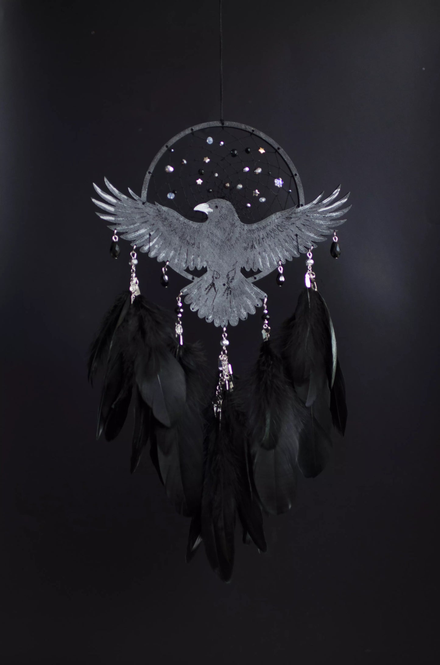 Handmade raven dream catcher with outstretched wings, set on a black hoop with a delicate web decorated with small reflective beads. Black feathers hang below with subtle crystal accents. Dark aesthetic wall hanging in charcoal and silver tones, detailed and symbolic design, perfect for moody interiors or meaningful gift.