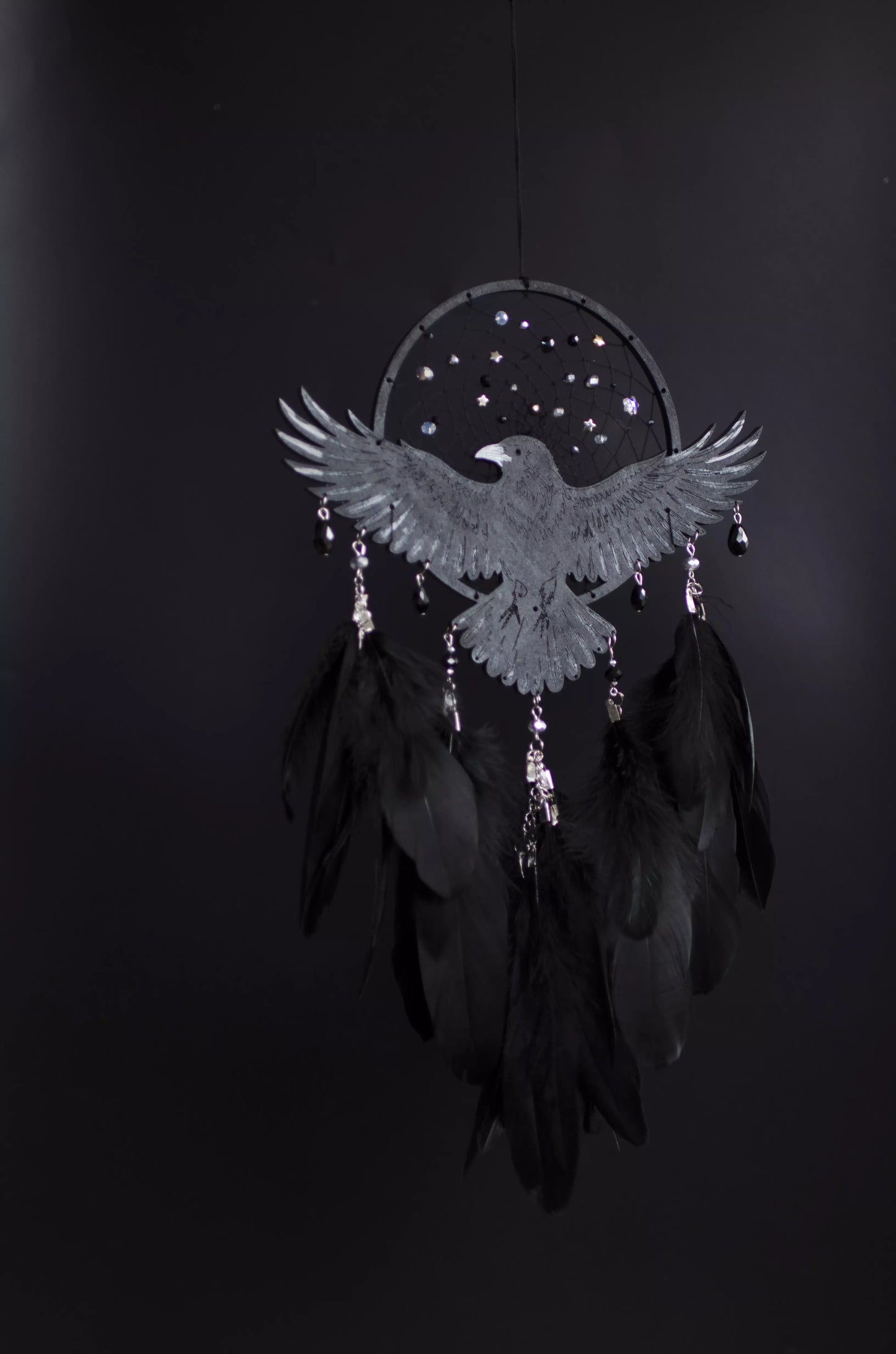 Handmade raven dream catcher with outstretched wings, set on a black hoop with a delicate web decorated with small reflective beads. Black feathers hang below with subtle crystal accents. Dark aesthetic wall hanging in charcoal and silver tones, detailed and symbolic design, perfect for moody interiors or meaningful gift.