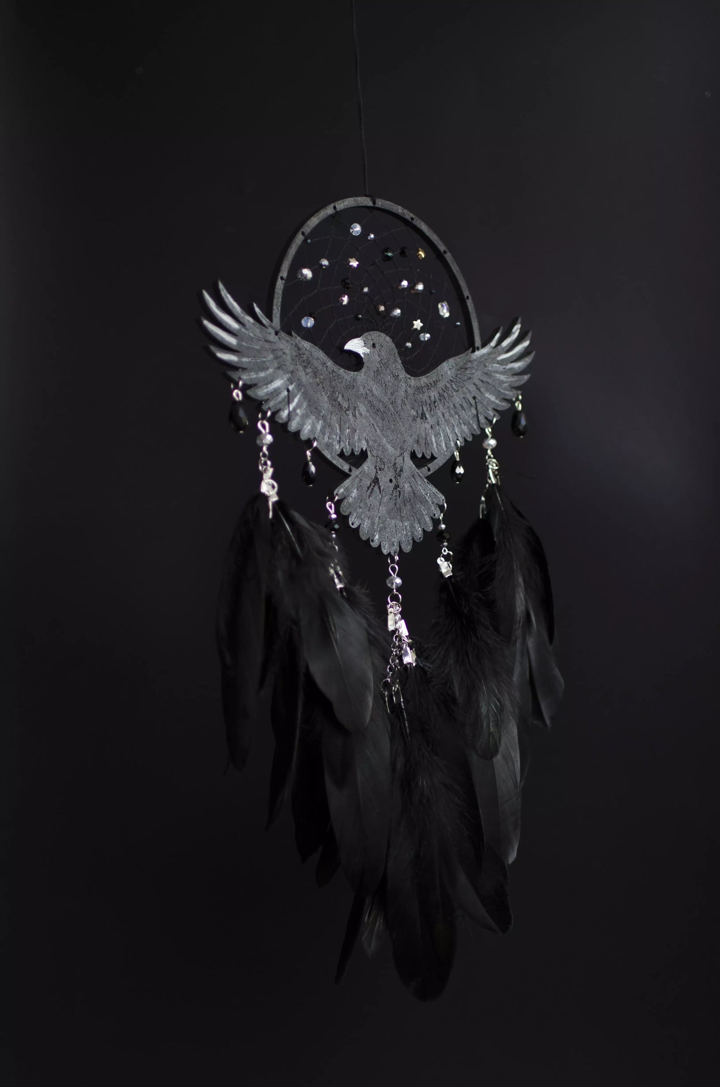 Handmade raven dream catcher with outstretched wings, set on a black hoop with a delicate web decorated with small reflective beads. Black feathers hang below with subtle crystal accents. Dark aesthetic wall hanging in charcoal and silver tones, detailed and symbolic design, perfect for moody interiors or meaningful gift.