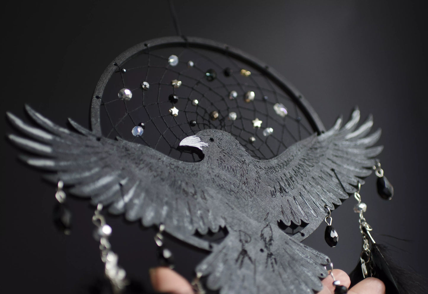 Handmade raven dream catcher with outstretched wings, set on a black hoop with a delicate web decorated with small reflective beads. Black feathers hang below with subtle crystal accents. Dark aesthetic wall hanging in charcoal and silver tones, detailed and symbolic design, perfect for moody interiors or meaningful gift.