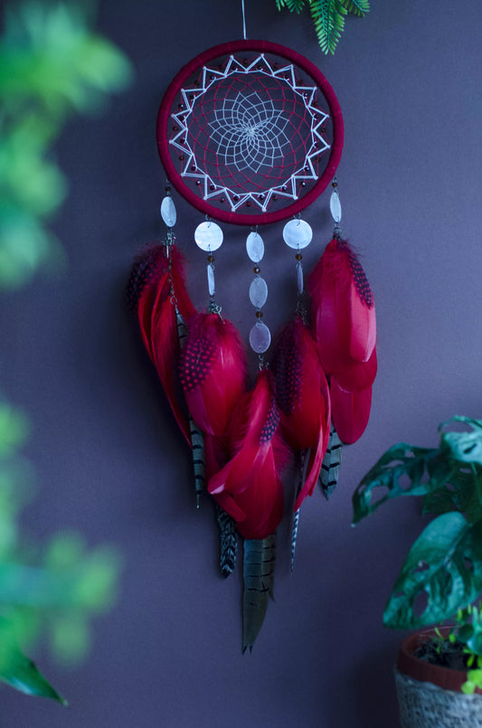 Red dream catcher with a detailed white web, adorned with white beads and vibrant red feathers. Perfect for adding a touch of boho charm and positive energy to your space. Hangs elegantly against a backdrop of greenery, creating a striking visual contrast.