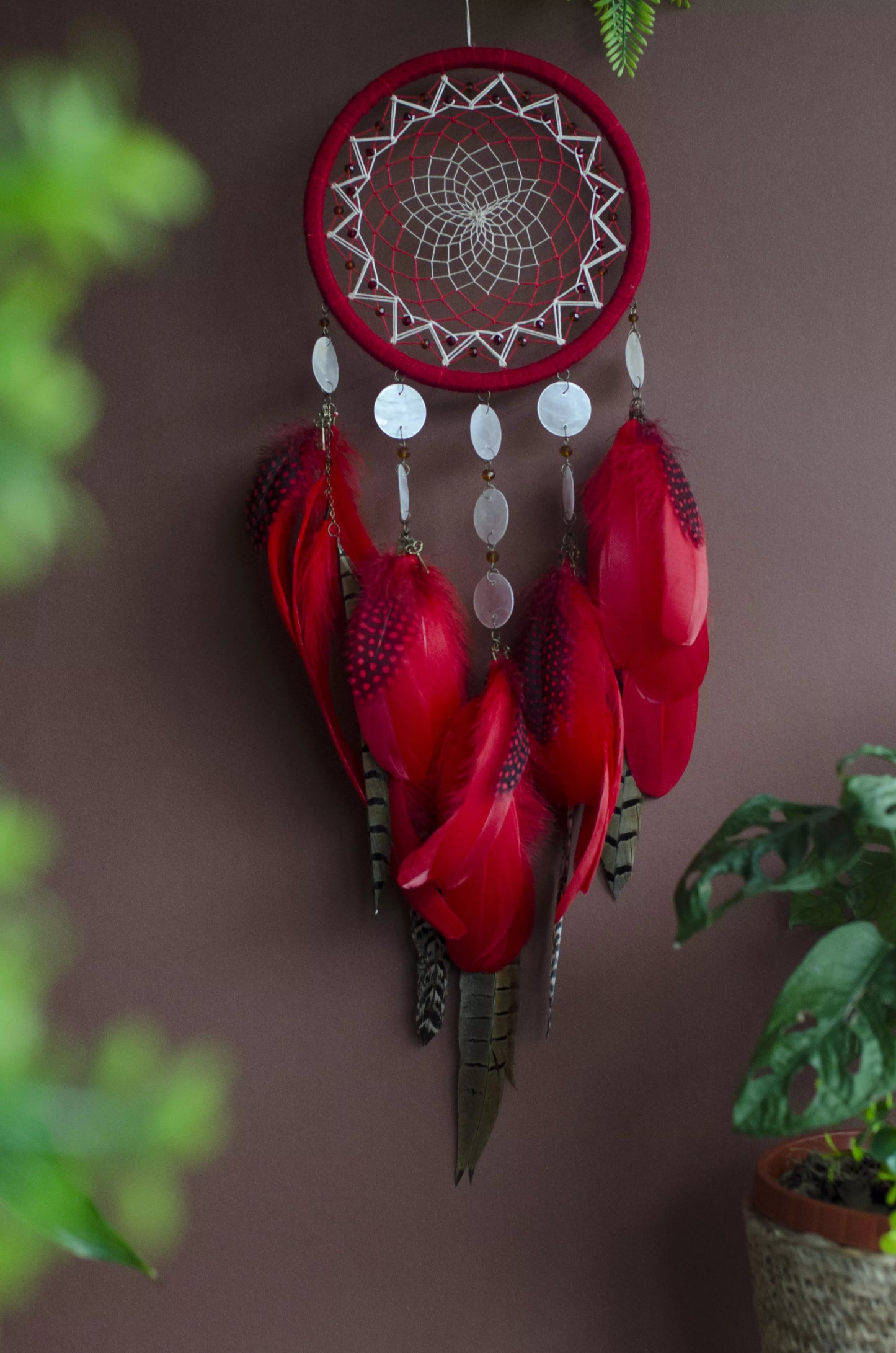 Red dream catcher with a detailed white web, adorned with white beads and vibrant red feathers. Perfect for adding a touch of boho charm and positive energy to your space. Hangs elegantly against a backdrop of greenery, creating a striking visual contrast.