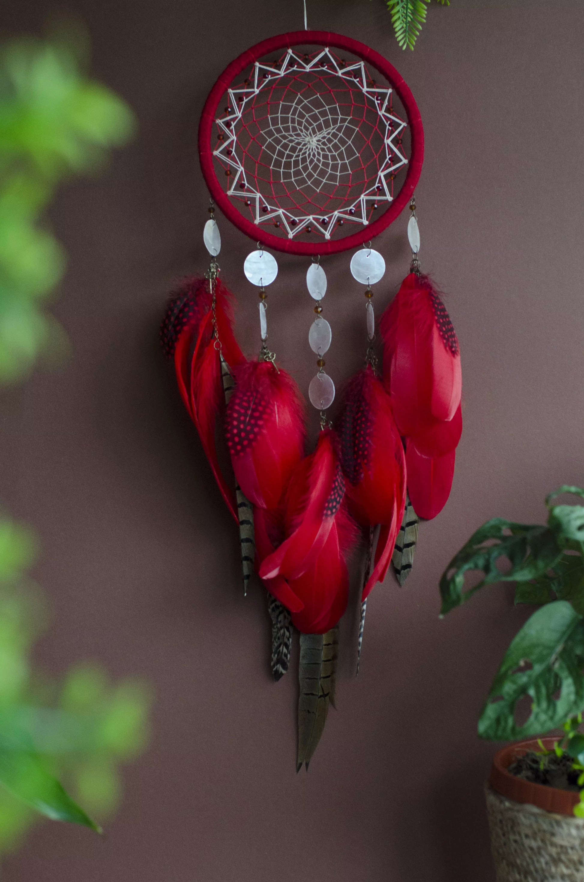 Red dream catcher with a detailed white web, adorned with white beads and vibrant red feathers. Perfect for adding a touch of boho charm and positive energy to your space. Hangs elegantly against a backdrop of greenery, creating a striking visual contrast.