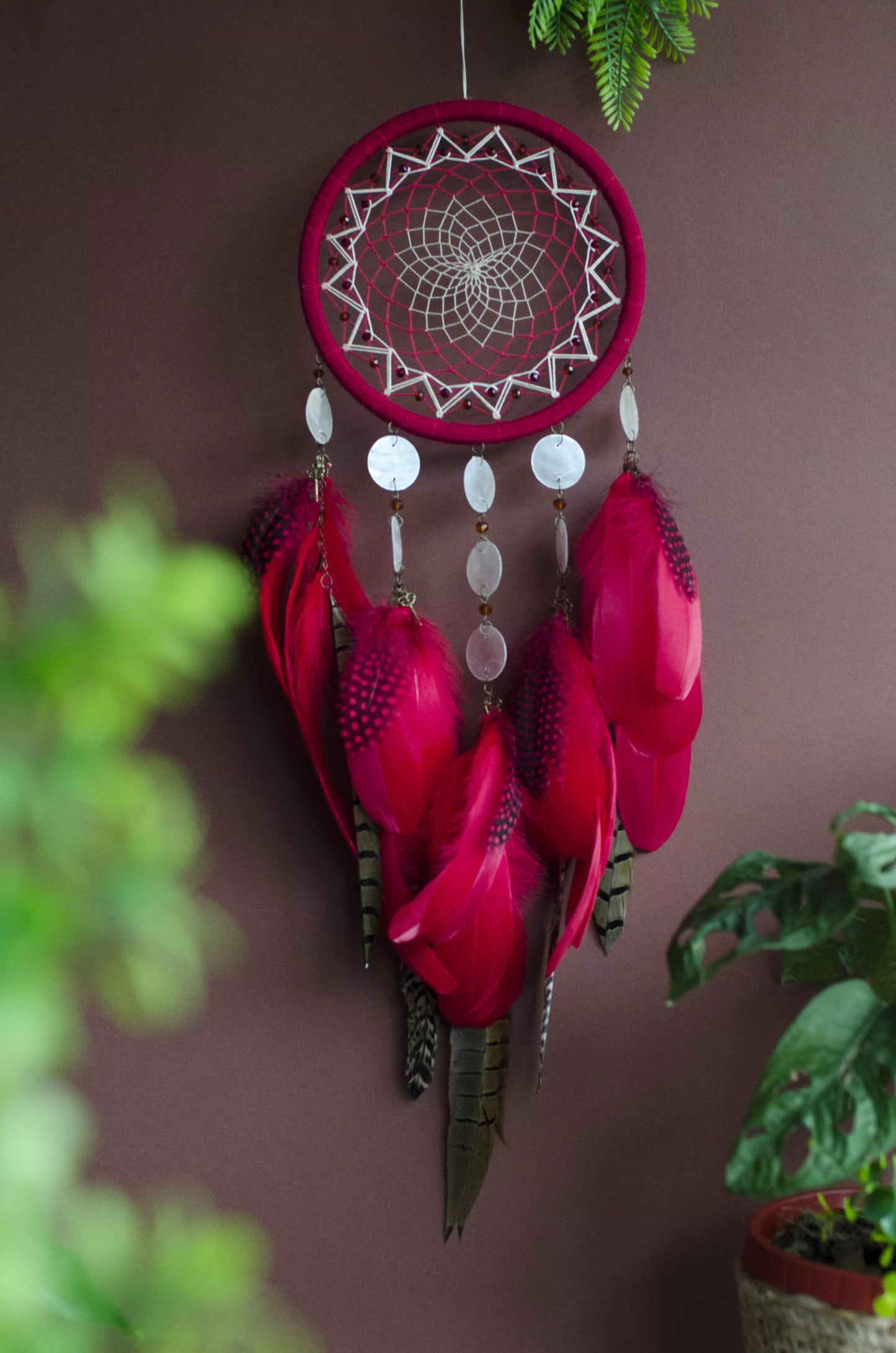 Red dream catcher with a detailed white web, adorned with white beads and vibrant red feathers. Perfect for adding a touch of boho charm and positive energy to your space. Hangs elegantly against a backdrop of greenery, creating a striking visual contrast.