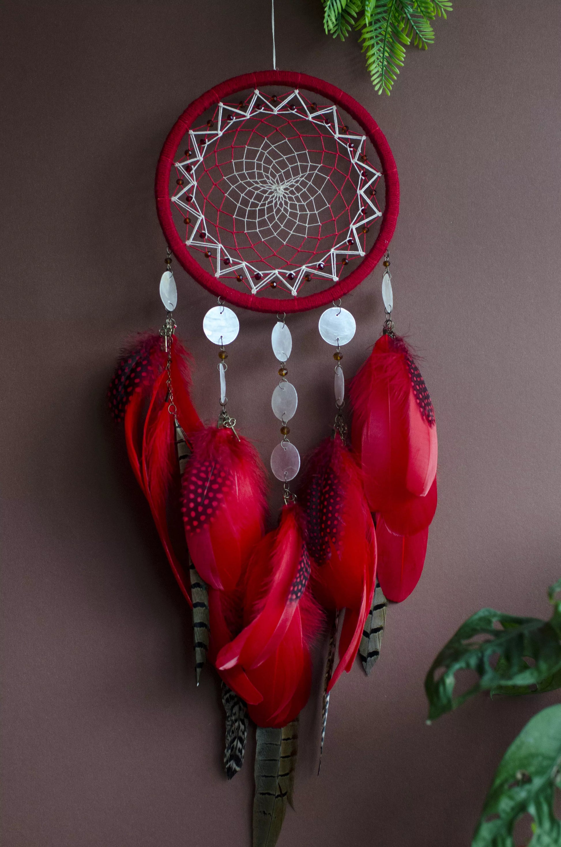 Red dream catcher with a detailed white web, adorned with white beads and vibrant red feathers. Perfect for adding a touch of boho charm and positive energy to your space. Hangs elegantly against a backdrop of greenery, creating a striking visual contrast.