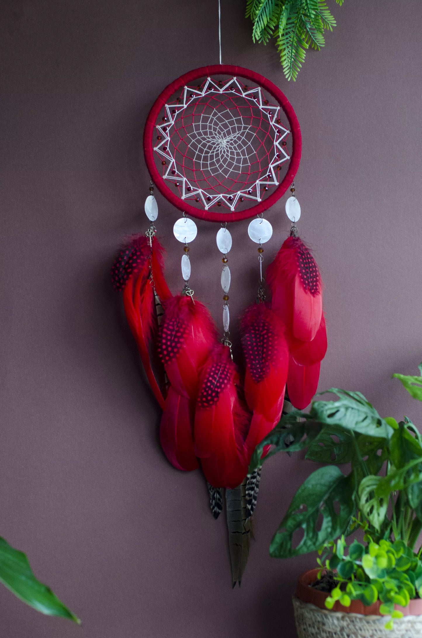 Red dream catcher with a detailed white web, adorned with white beads and vibrant red feathers. Perfect for adding a touch of boho charm and positive energy to your space. Hangs elegantly against a backdrop of greenery, creating a striking visual contrast.