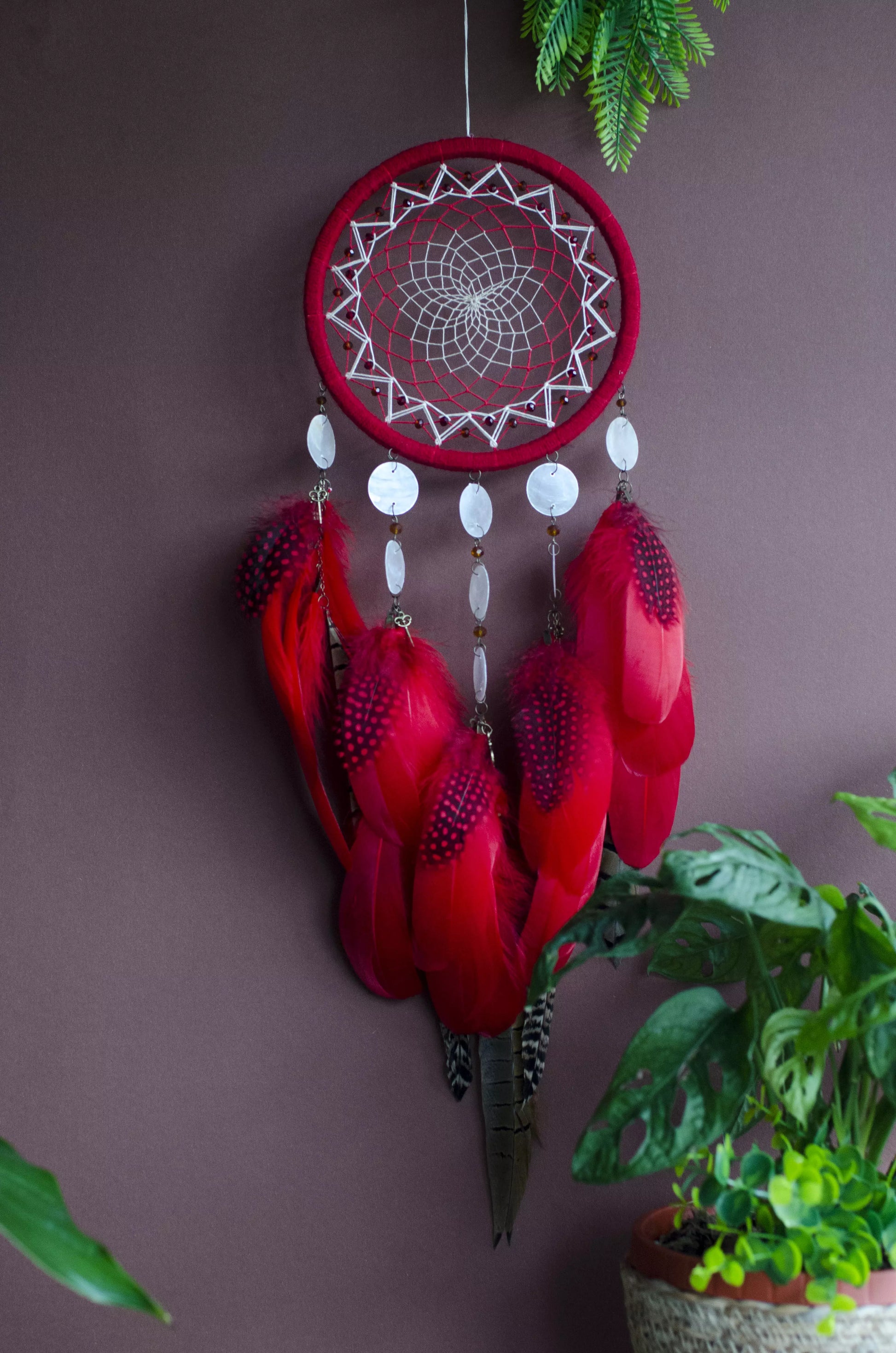 Red dream catcher with a detailed white web, adorned with white beads and vibrant red feathers. Perfect for adding a touch of boho charm and positive energy to your space. Hangs elegantly against a backdrop of greenery, creating a striking visual contrast.