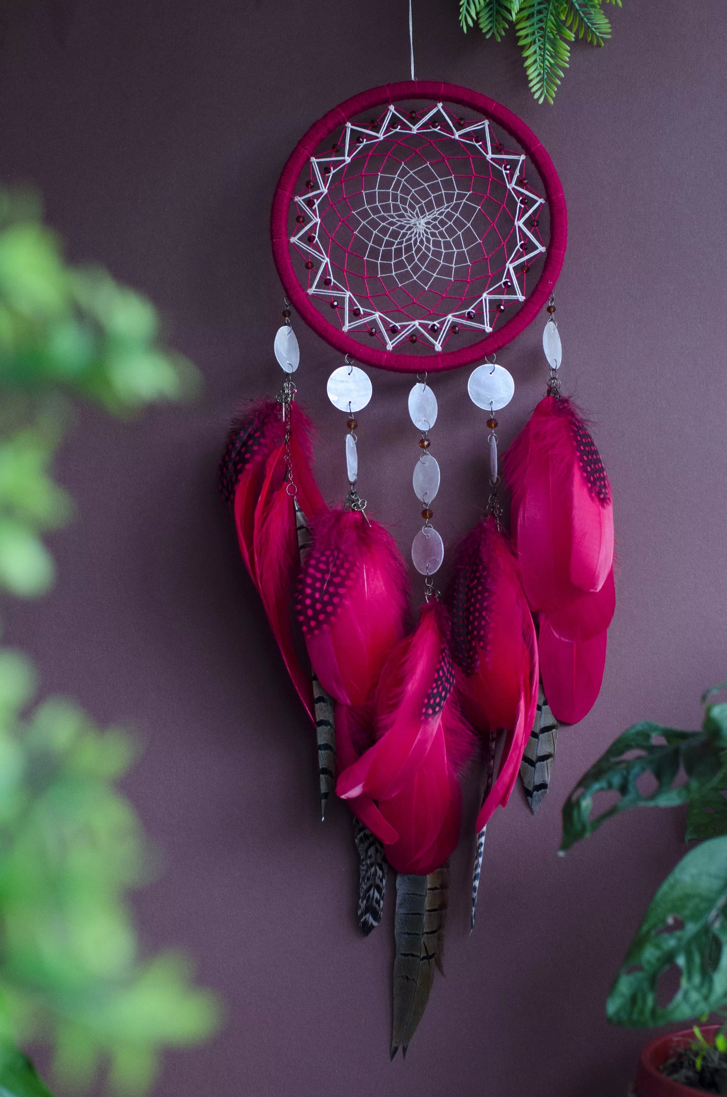 Red dream catcher with a detailed white web, adorned with white beads and vibrant red feathers. Perfect for adding a touch of boho charm and positive energy to your space. Hangs elegantly against a backdrop of greenery, creating a striking visual contrast.